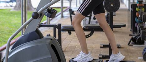 A person using an elliptical machine under a covered area with gym equipment in the background.