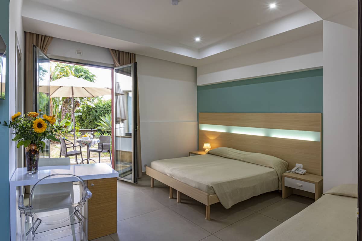 Une chambre d’hôtel avec un lit, une table de chevet et un bureau avec une chaise. Il y a une fenêtre avec des rideaux, un vase avec des fleurs et une vue sur l’extérieur.