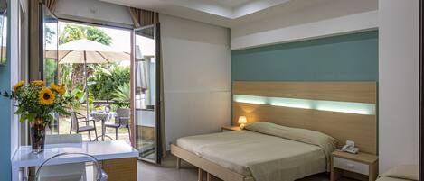 Habitación de hotel con cama, mesita de noche y escritorio con silla. Se observa una ventana con cortinas, un jarrón con flores y vistas al exterior.