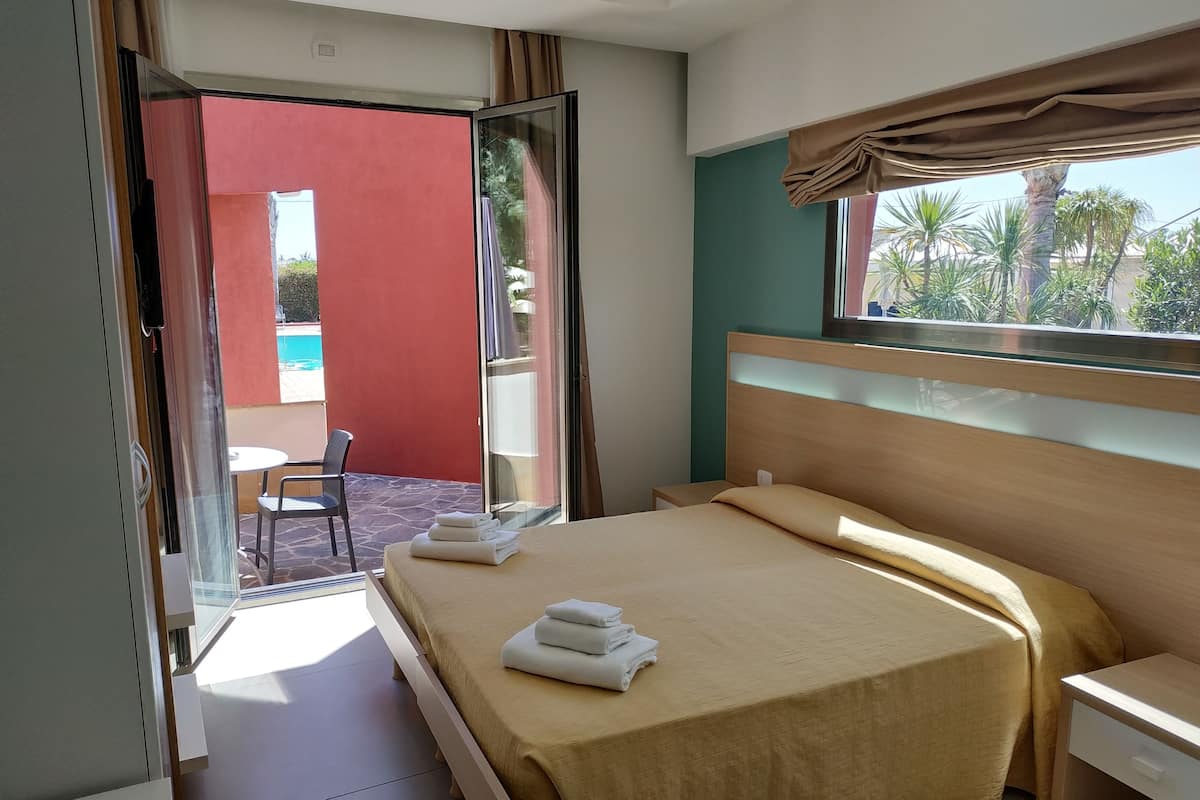 Une chambre d’hôtel comprenant un lit, une table de chevet, une fenêtre donnant sur des palmiers et une porte-fenêtre coulissante qui mène à un espace extérieur avec une piscine.
