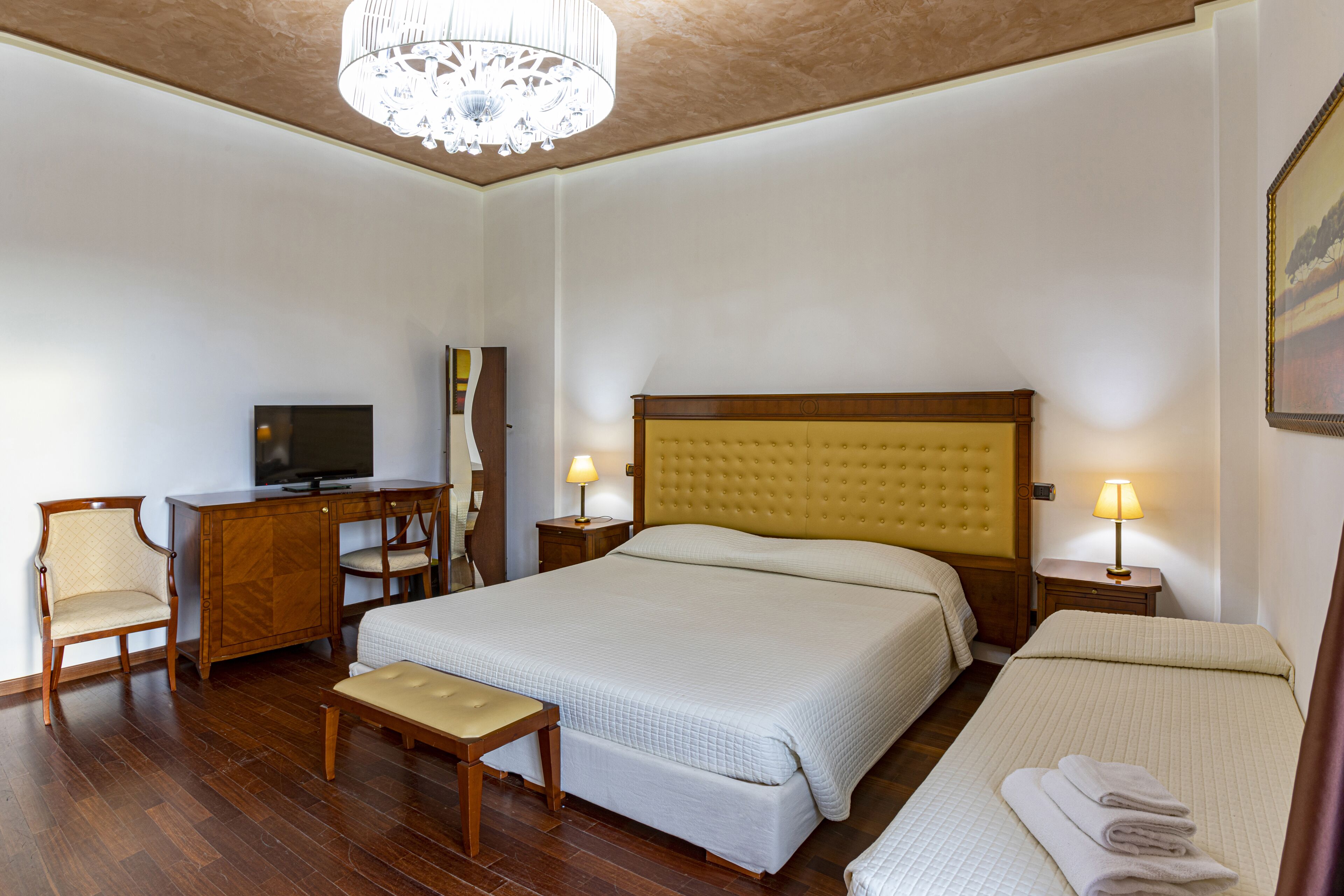 Una camera d'albergo con un letto grande, una scrivania con televisore, una sedia e due lampade ai comodini.