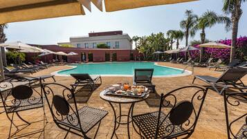 Zona junto a la piscina con sillones de descanso, una mesa con alimentos para el desayuno y un edificio al fondo.