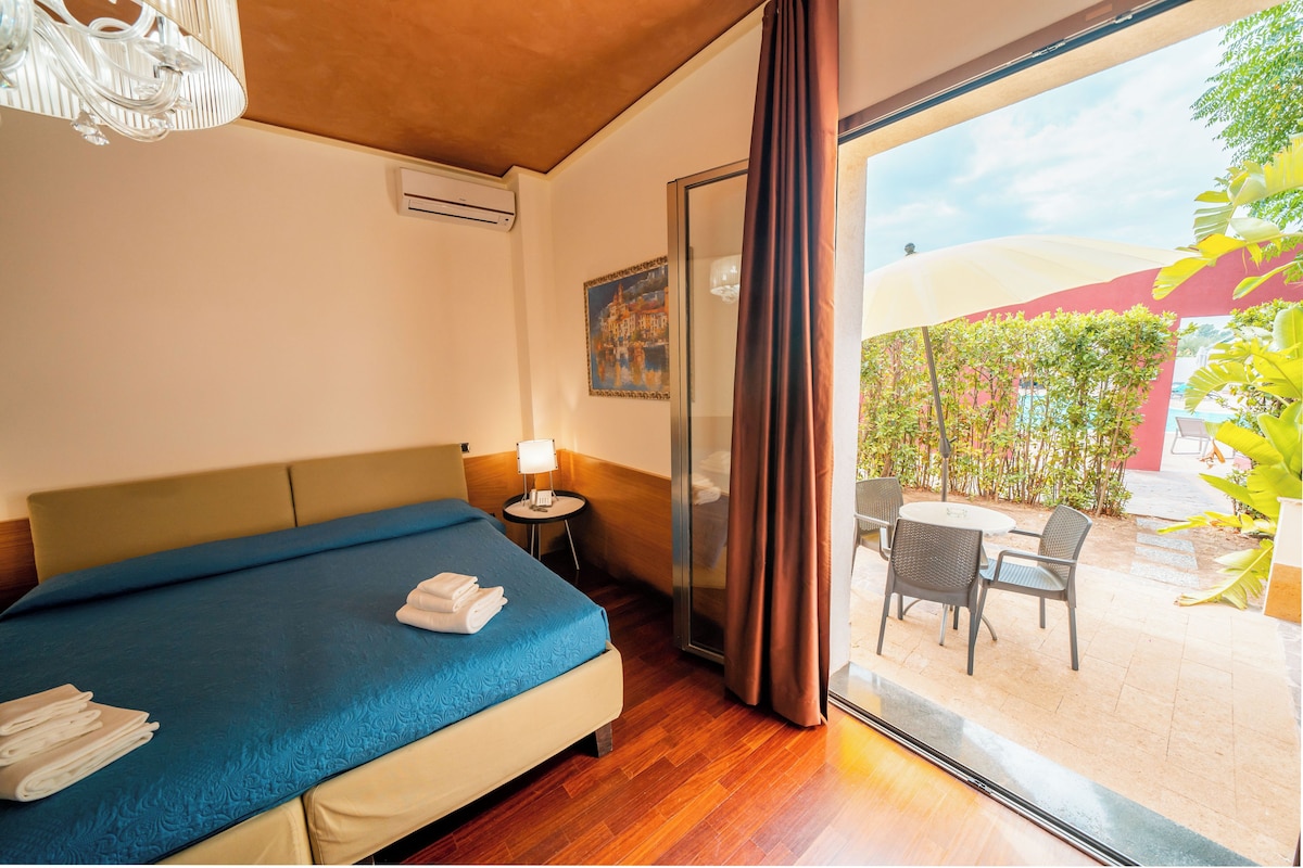 Una habitación de hotel con cama, una mesita con lámpara, ventana con cortinas y un área exterior con sillas y mesa.