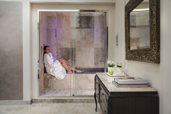 A person sitting in a glass-enclosed shower stall with a marble floor, a white robe, and a small table with a plant and a book.