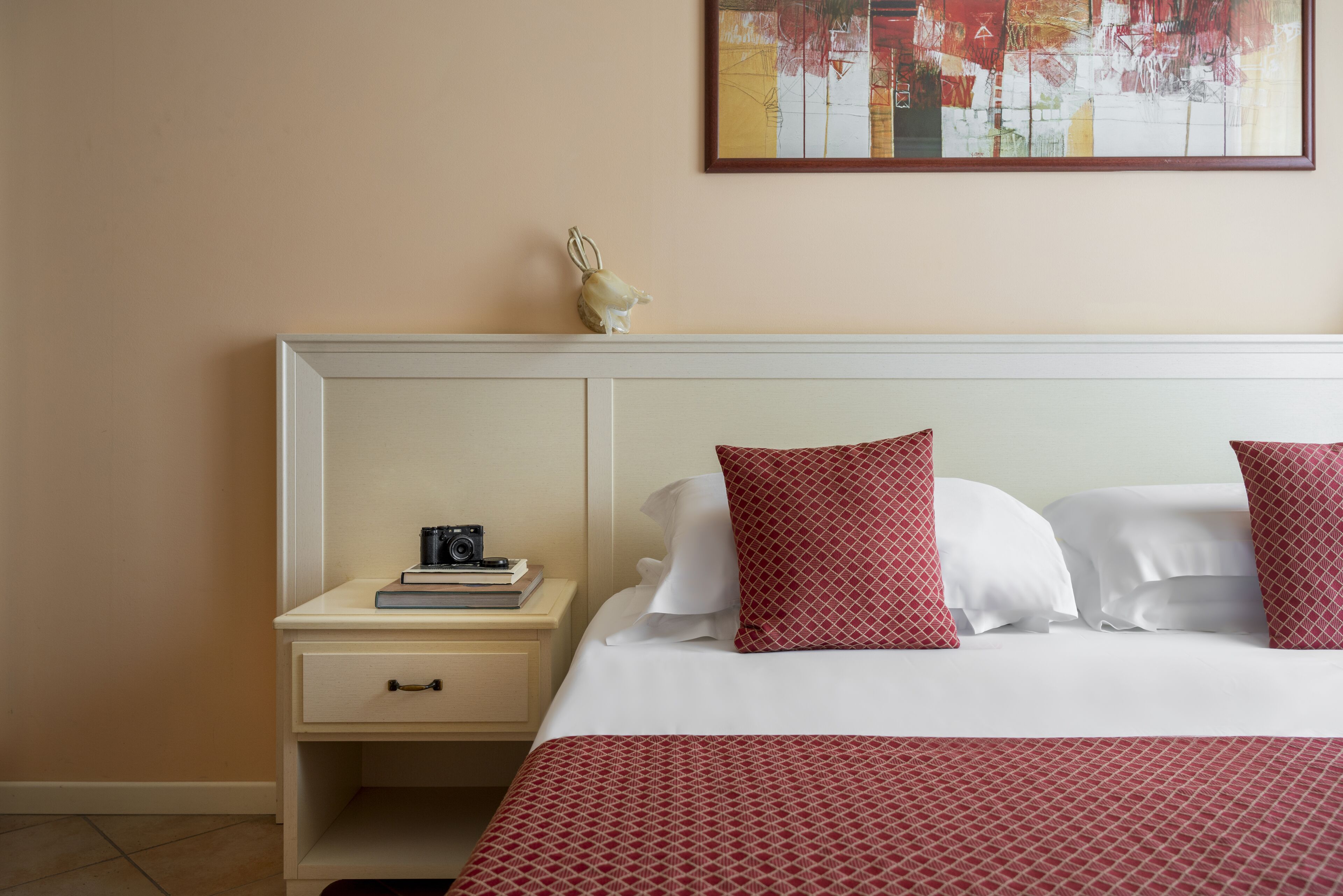 A hotel room with a bed, a nightstand, a camera, and a framed painting on the wall.
