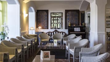 A spacious lounge area with wicker chairs arranged around a central table, a piano, and a bookshelf.