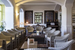 A spacious lounge area with wicker chairs arranged around a central table, a piano, and a bookshelf.
