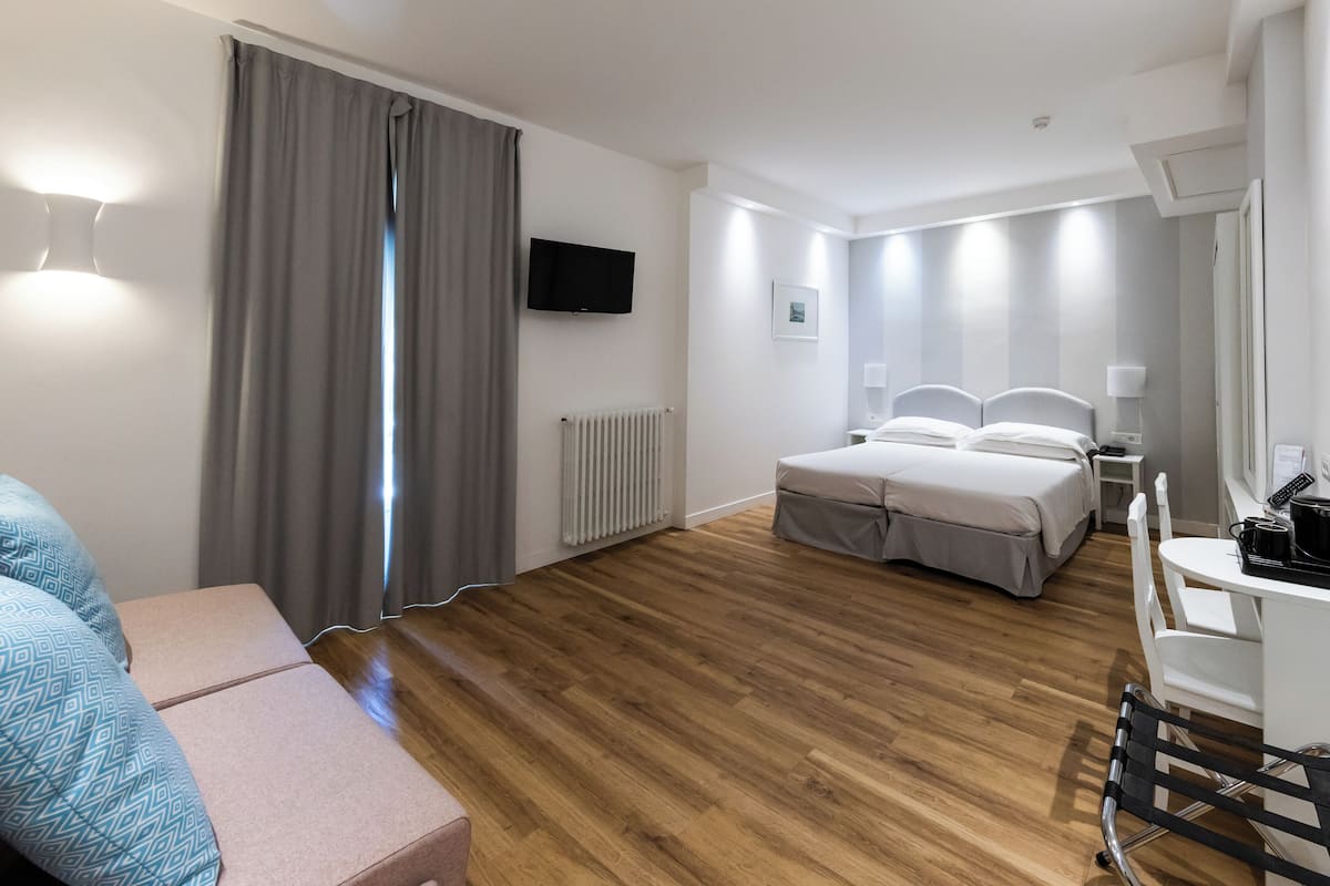 Ein modernes Hotelzimmer mit Holzboden, einem Bett mit weißer Bettwäsche, einem Schreibtisch mit Stuhl, einem an der Wand montierten Fernseher und einem Fenster mit Vorhängen.