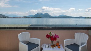 Un balcón con dos sillones blancos de mimbre, una mesa con un jarrón de flores y vistas a un lago con montañas al fondo.