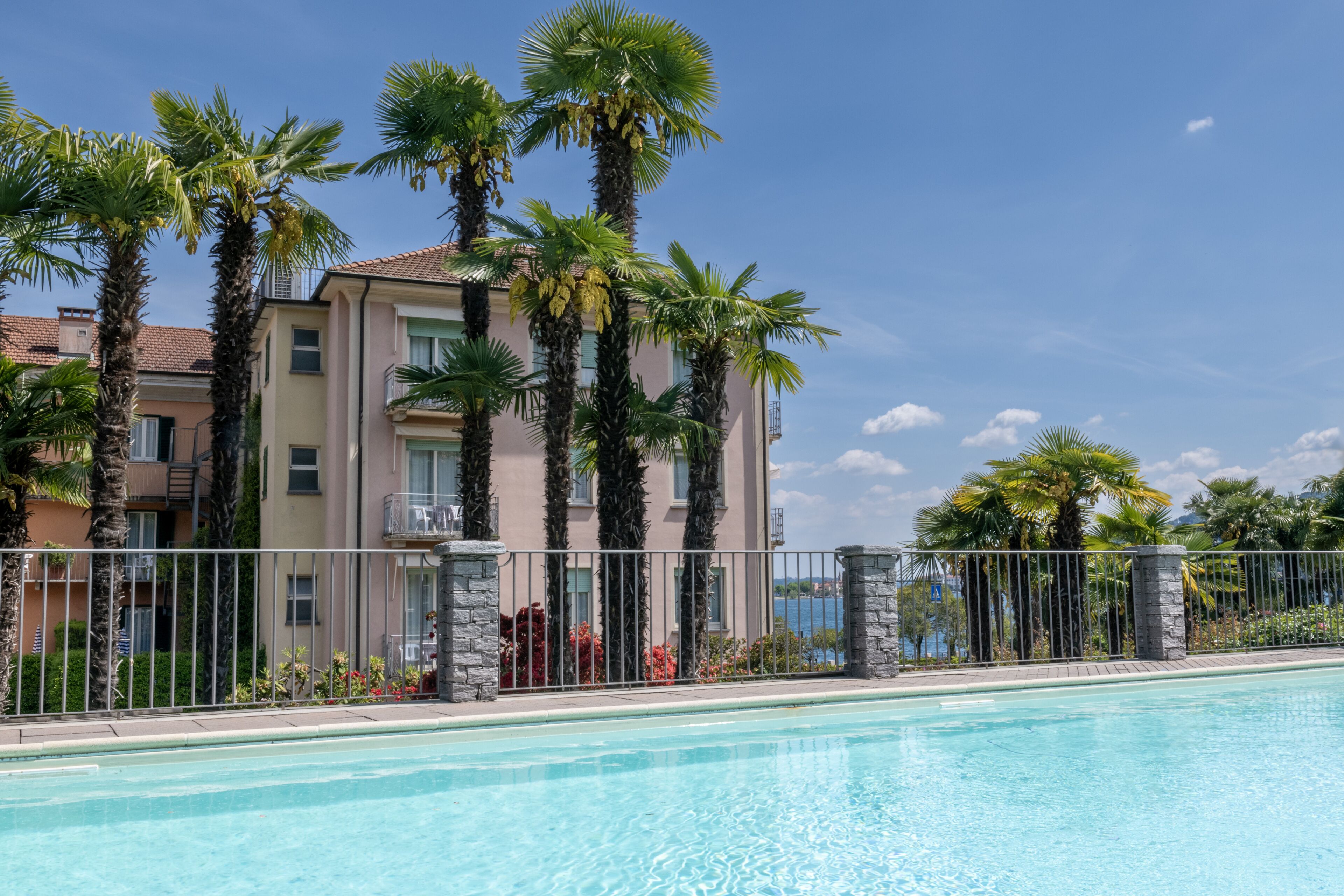 Una piscina con agua azul cristalina, rodeada por una cerca de metal, palmeras y un edificio con balcones.