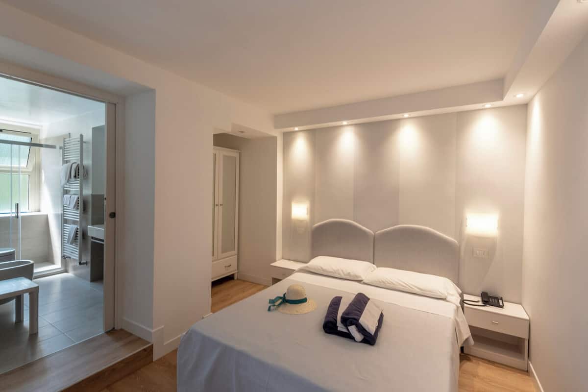 Ein modernes Schlafzimmer mit einem Bett, Nachttischen und einem kleinen Tisch neben dem Bett. In der Ecke steht eine Bank und eine Tür führt ins Badezimmer.
