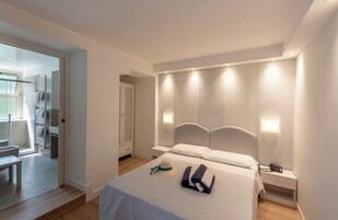 Un dormitorio moderno con cama, mesitas de noche y un banco en la esquina. Se ve una puerta que da a un baño.