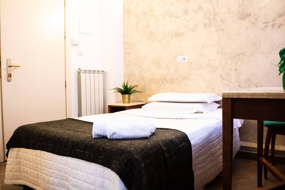 A neatly made bed with a black and white quilt, a small bedside table with a plant, and a wooden chair.
