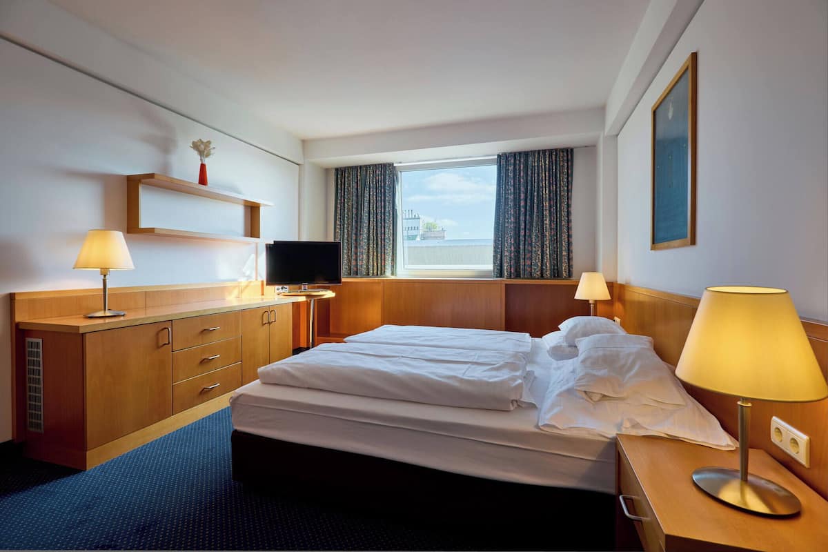 A hotel room with a bed, two bedside lamps, a TV, a wooden dresser, and a window with curtains.