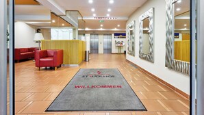 A hotel reception area with a reception desk, red seating, a carpet with 'STUDHOLF' and 'WILLKOMMEN' text, and a mirror on the wall. - Hotel Strudlhof (Vienna)