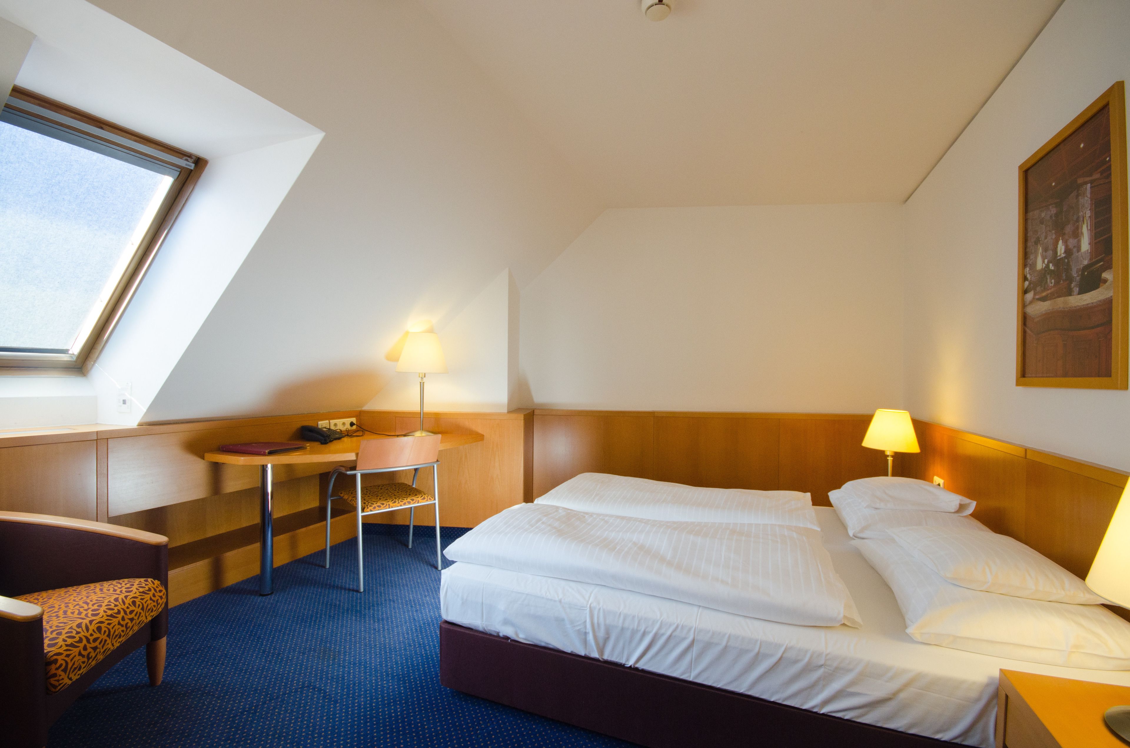 A hotel room with a bed, two bedside lamps, a desk with a chair, and a window with blinds.