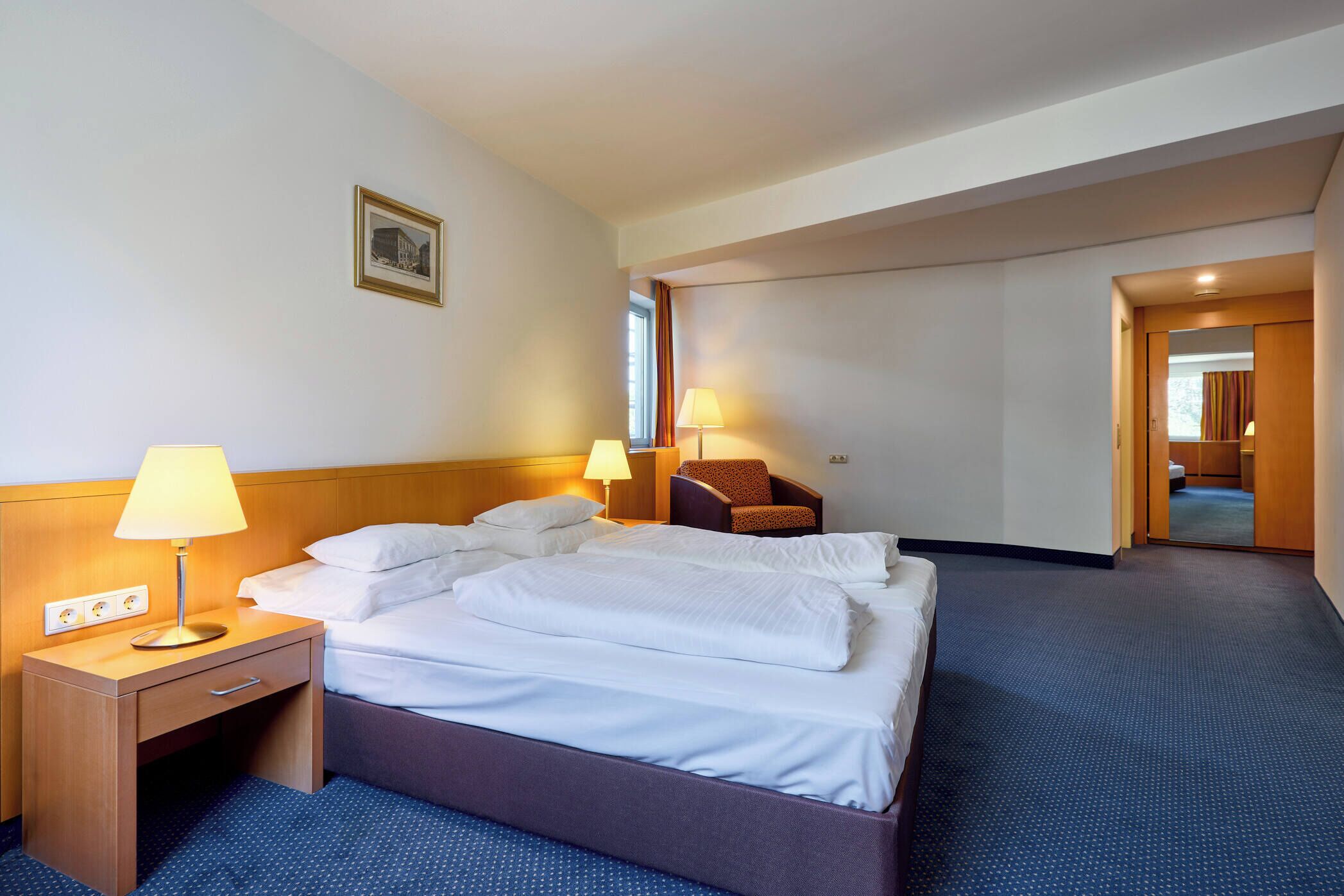 Una habitación de hotel con una cama, lámparas de noche, una silla y una puerta que da a un baño.