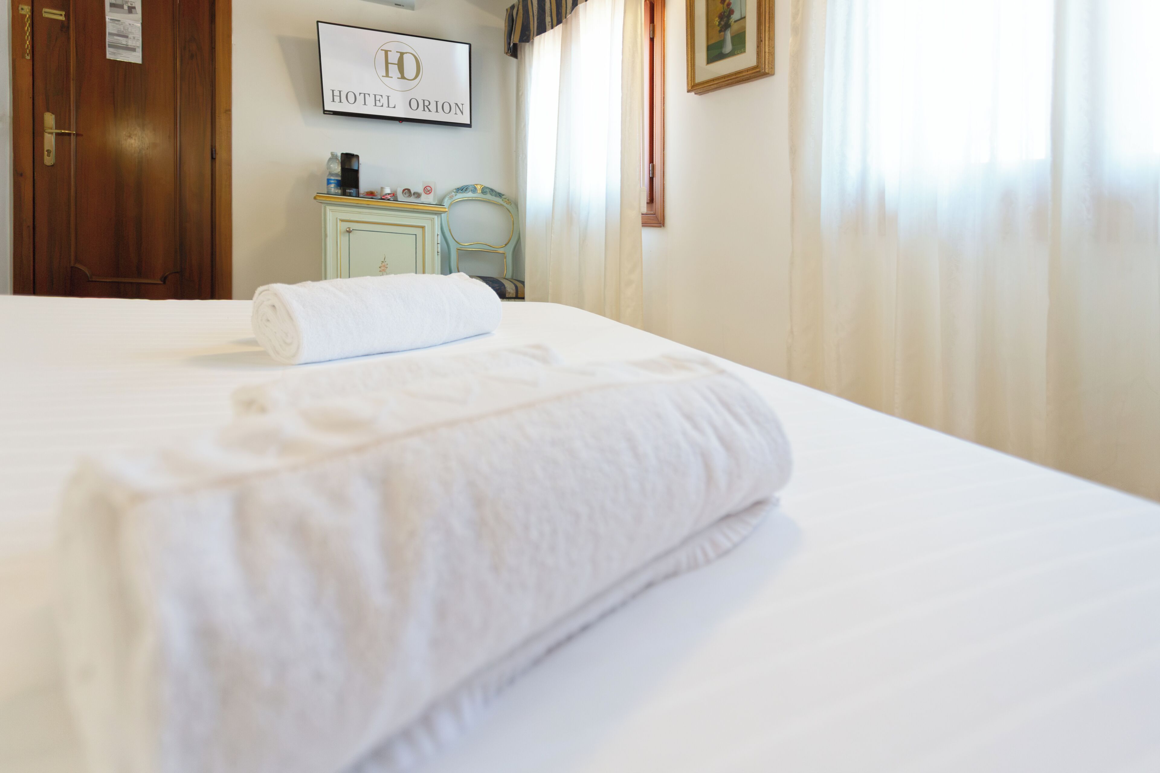 A neatly made bed with a rolled white towel, a framed sign reading 'HOTEL ORION' on the wall, and a wooden door.