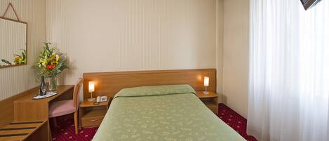 A hotel room with a bed, a desk with a chair, a telephone, a lamp, and a TV on the wall.