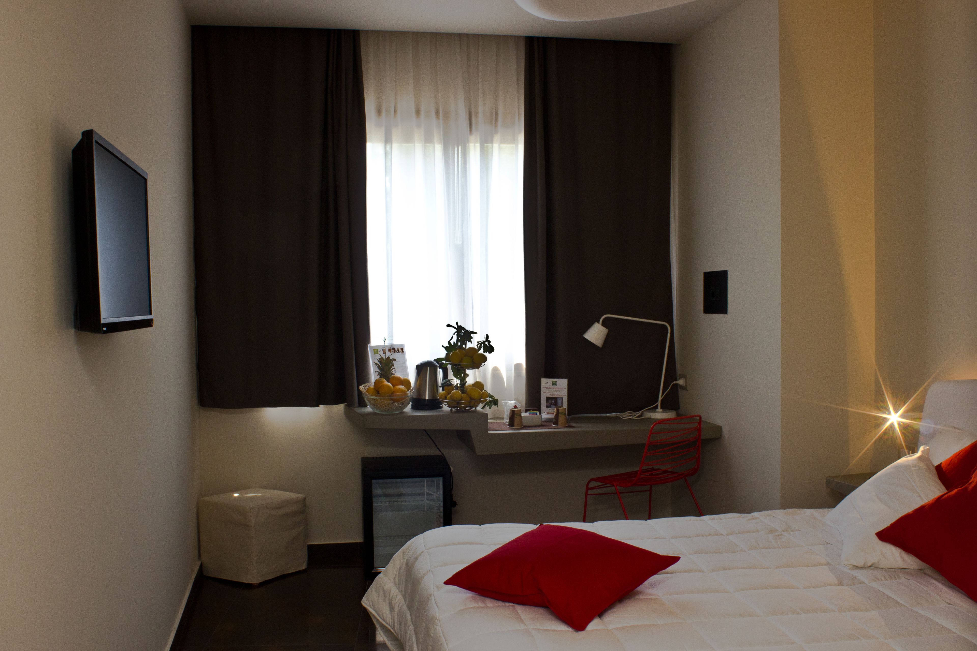 A modern hotel room with a bed, a desk with a chair, a small trash bin, and a window with curtains.