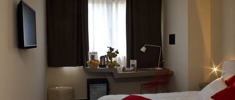 A modern hotel room with a bed, a desk with a chair, a small trash bin, and a window with curtains.