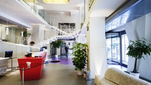 A modern hotel lobby with a red reception desk, a staircase, and potted plants.