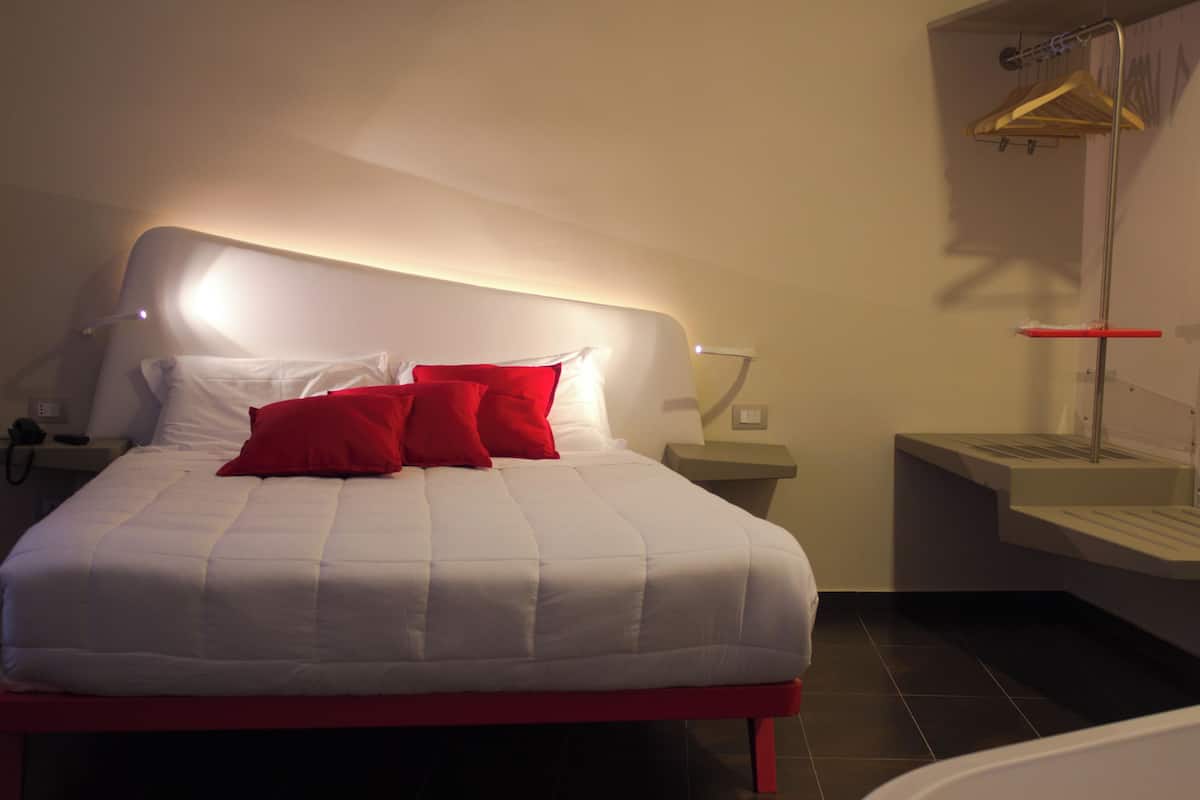 A modern bedroom with a bed, red pillows, a bedside table, a nightstand, and a wall-mounted shelf.