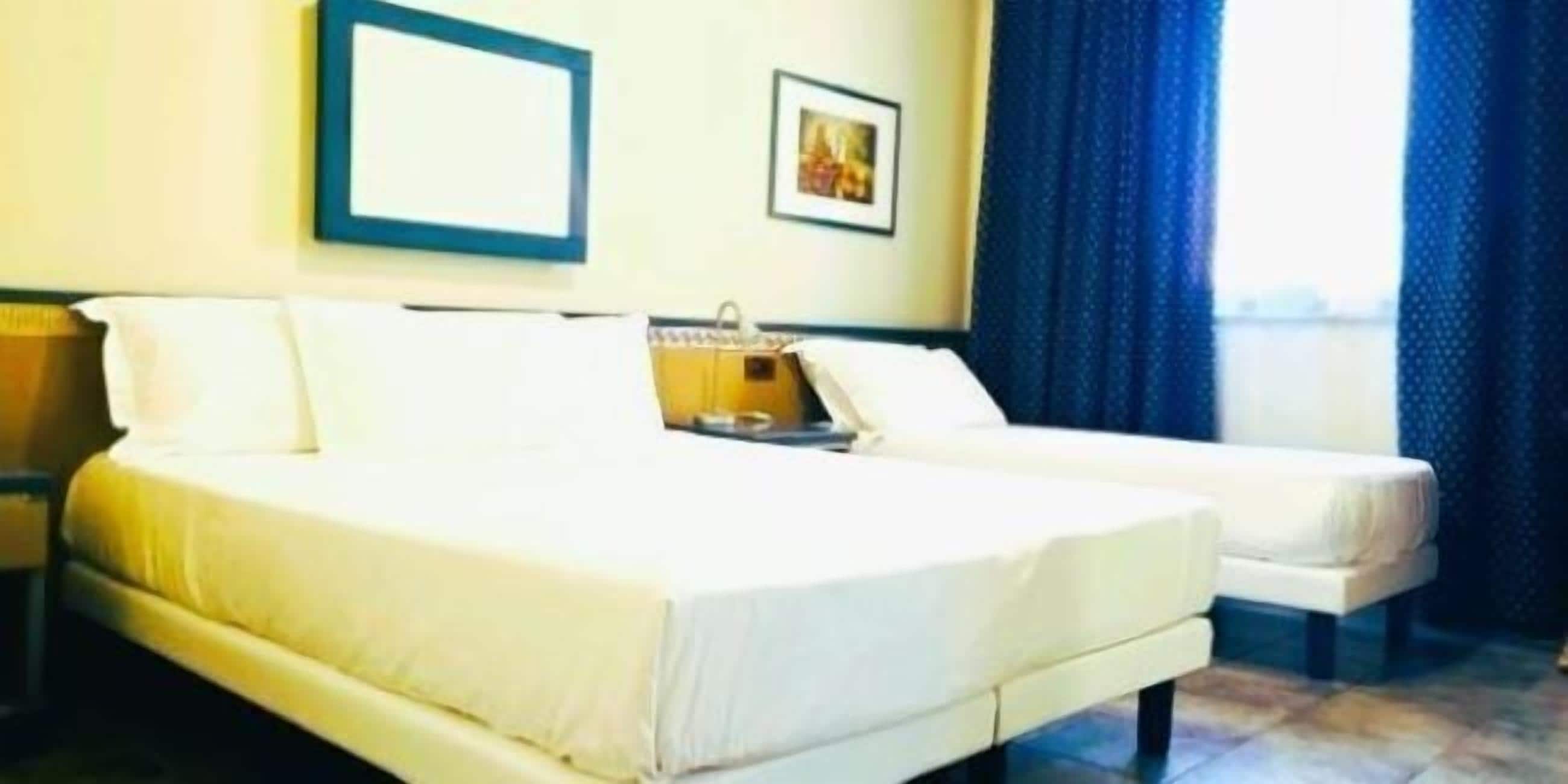 Hotellihuone, jossa on kaksi sänkyä, yöpöytä, seinällä oleva kehystetty kuva ja ikkuna, jossa on verhot.