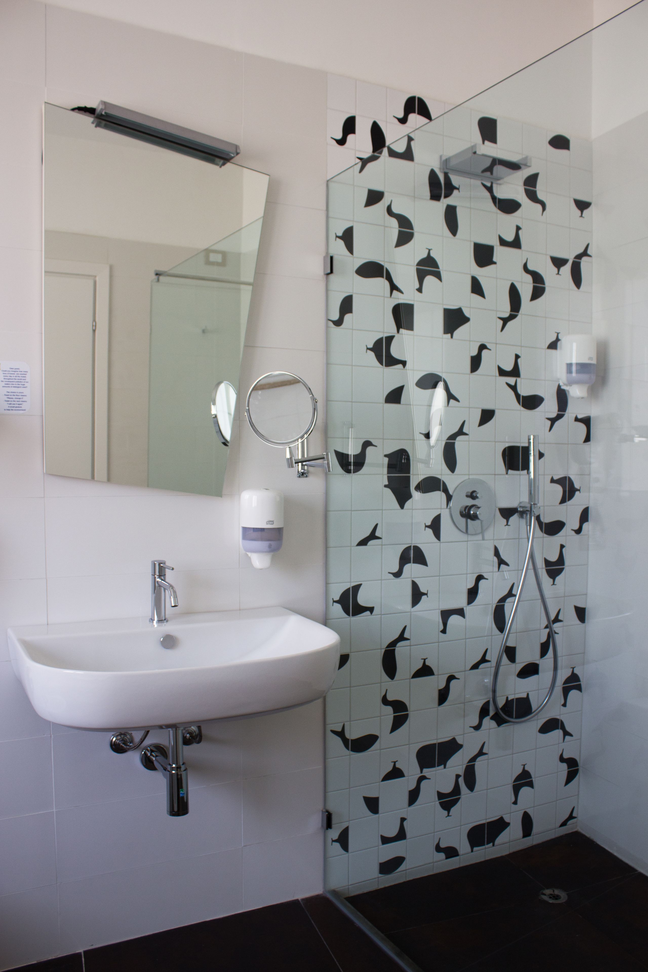 A bathroom with a white sink, a mirror, and a glass shower enclosure with black fish tiles.