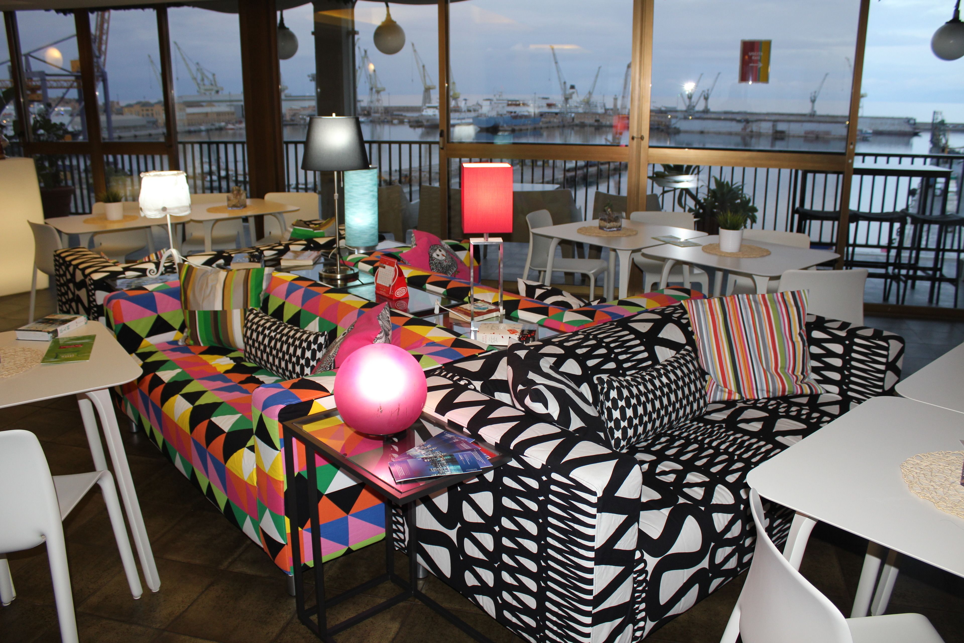 Ein moderner Lounge-Bereich mit einem farbenfrohen, geometrischen Sofa, weißen Tischen und Blick auf den Hafen.