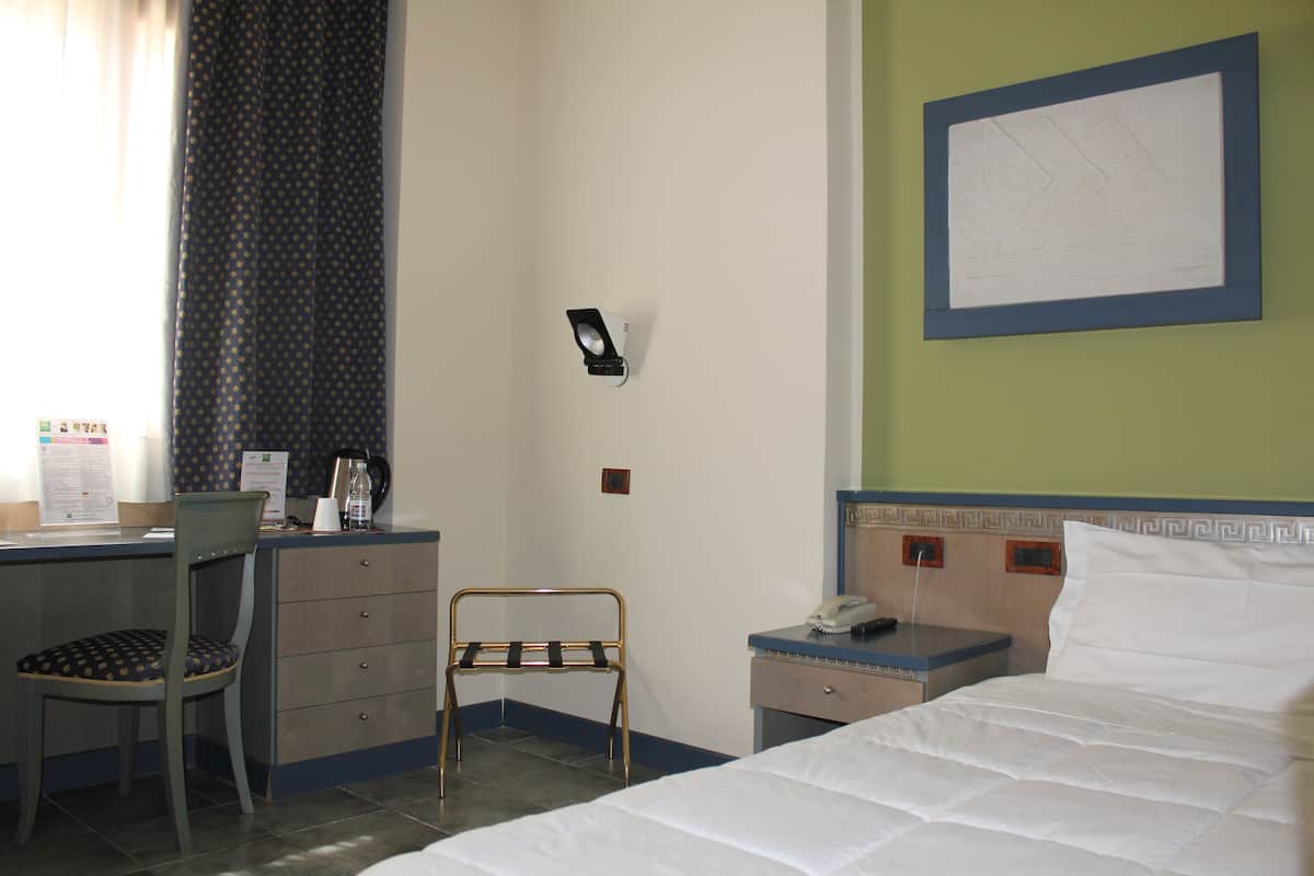 A hotel room with a bed, a desk with a chair, a small table with a phone, and a wall-mounted light.