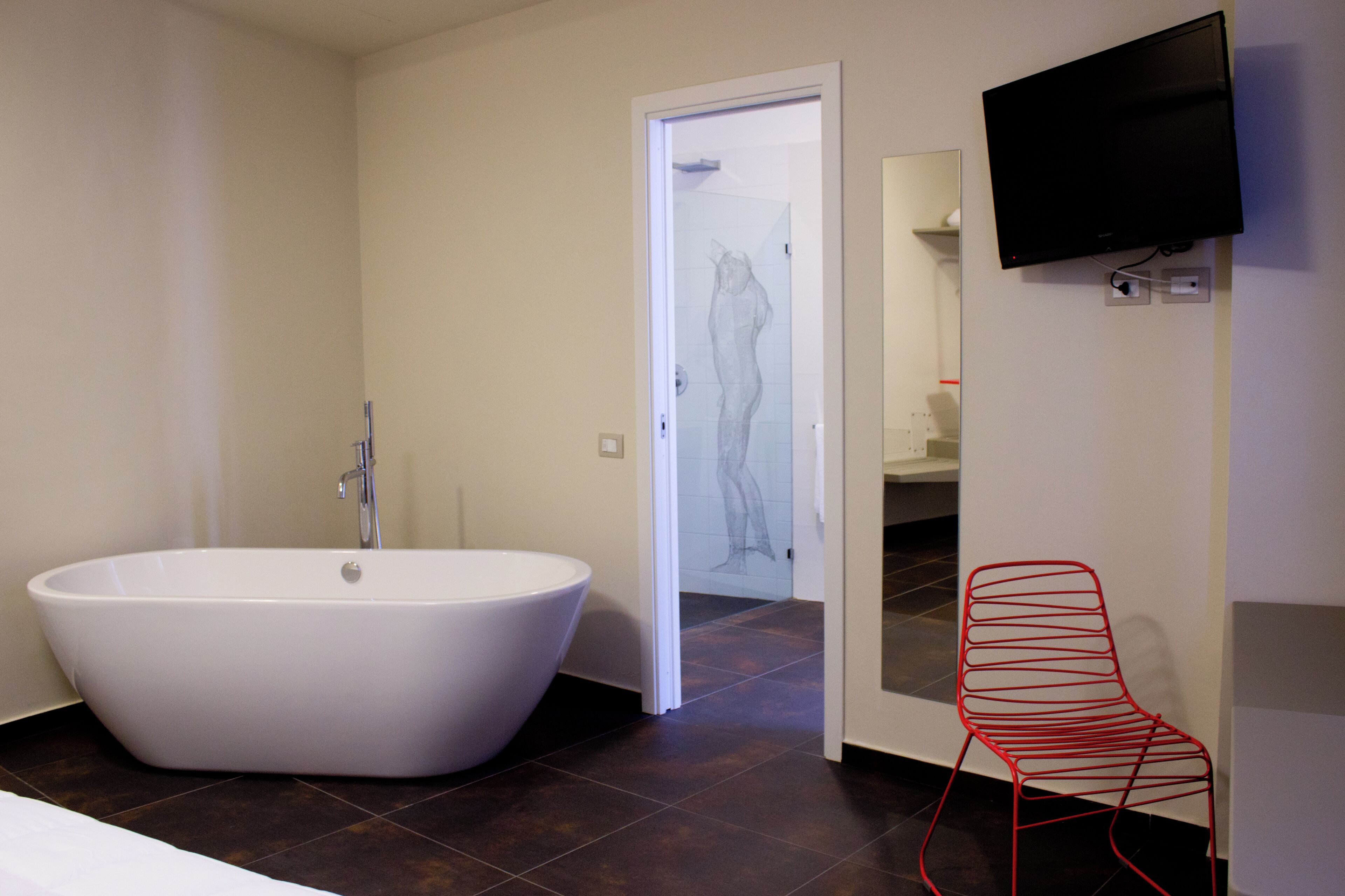 A modern bathroom with a freestanding bathtub, a red chair, and a flat-screen TV mounted on the wall.