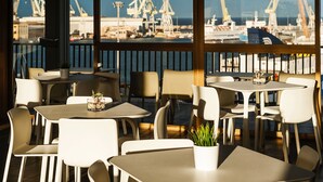 A restaurant with outdoor seating, white chairs, and tables, overlooking a harbor with cranes and boats.