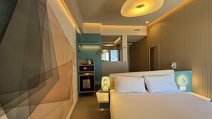 A modern hotel room with a large bed, a small kitchenette, and a bathroom visible through an open door.