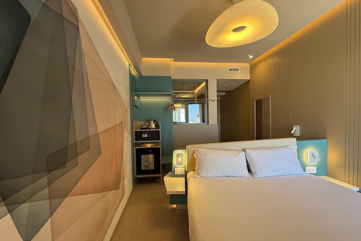 A modern hotel room with a large bed, a small kitchenette, and a bathroom visible through an open door.