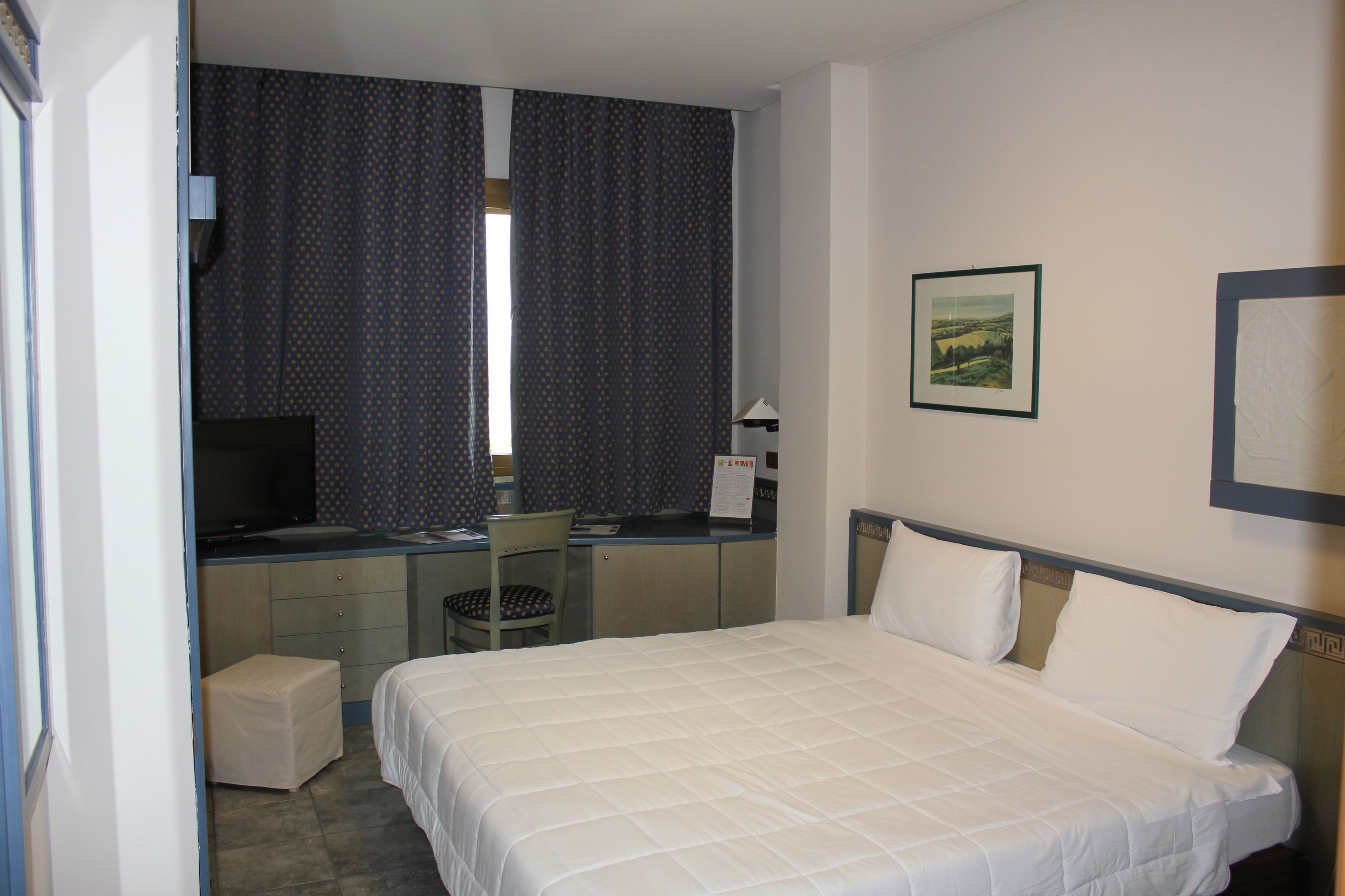Quarto de hotel com cama, escrivaninha, cadeira, televisão e janela com cortinas.
