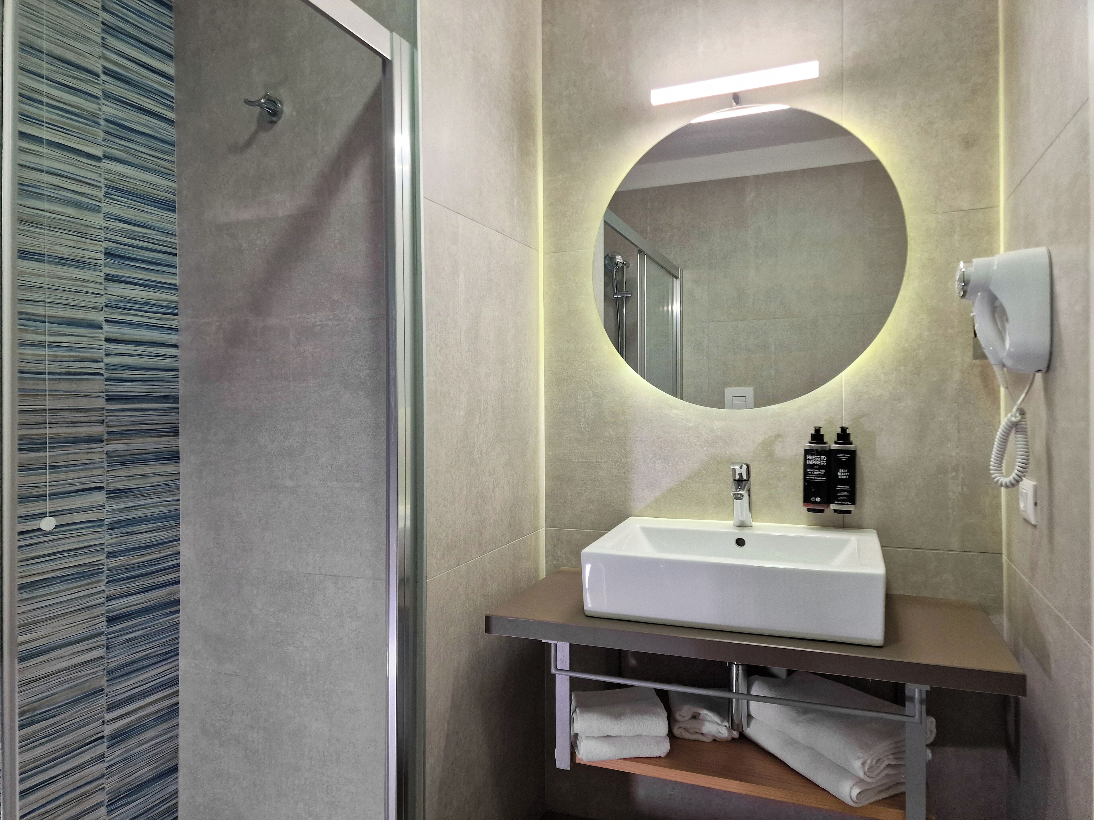 Un baño moderno con un lavabo blanco, un espejo redondo, un cabezal de ducha manual y un perchero para toallas.