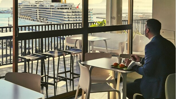 A man dining at a table with a view of a cruise ship and a building labeled "USCITA ELN" in the background.