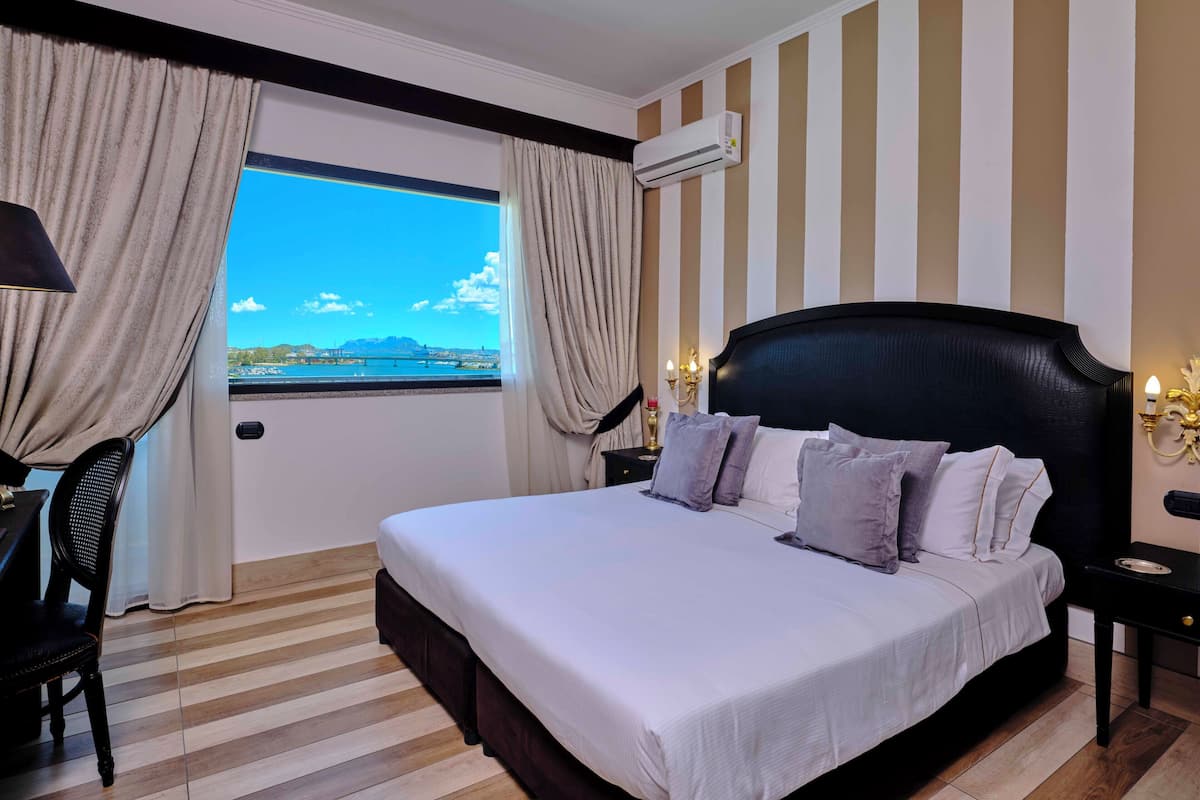 A hotel room with a large bed, a desk, a chair, an air conditioning unit, and a view of a body of water.