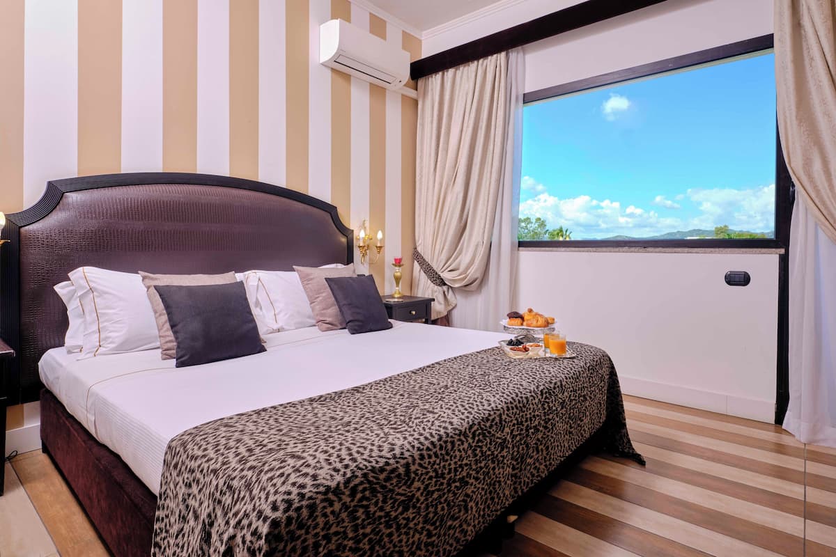 A hotel room with a large bed, striped walls, an air conditioning unit, a nightstand with a lamp, and a breakfast tray on the windowsill.