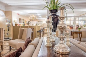 A well-lit, elegant interior with a seating area featuring a sofa and armchairs, a wooden table with decorative vases, and a bar area in the background.