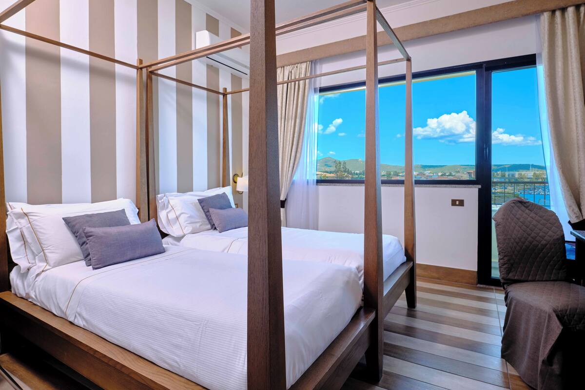 A four-poster bed with white linens and gray pillows, a wooden floor, a view of the sea through a large window, and a balcony with a chair.