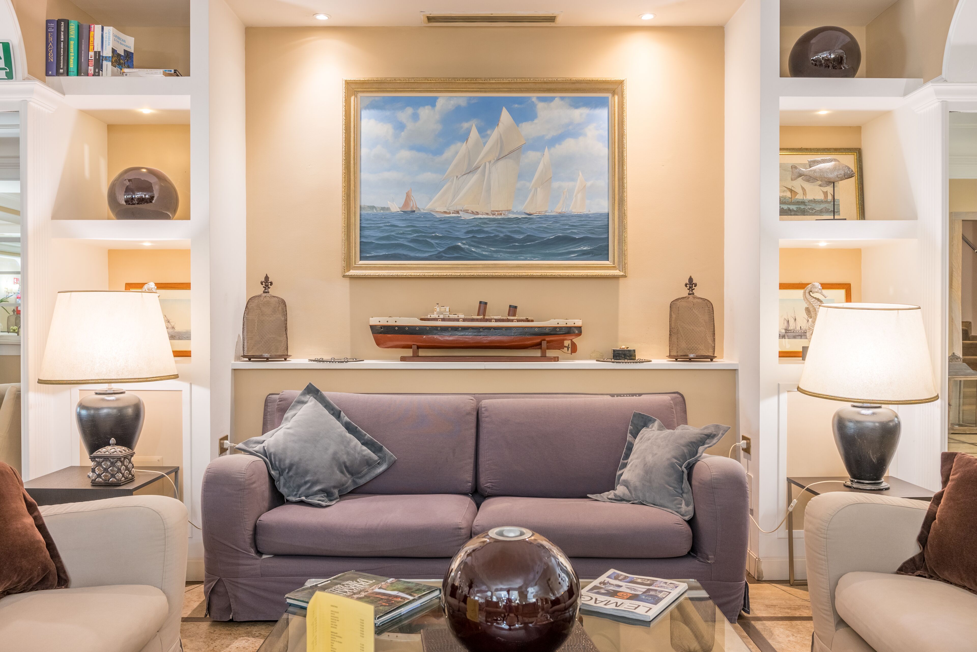 A living room with a sofa, two side tables with lamps, a coffee table, a painting of sailboats, and a bookshelf with books and decorative items.