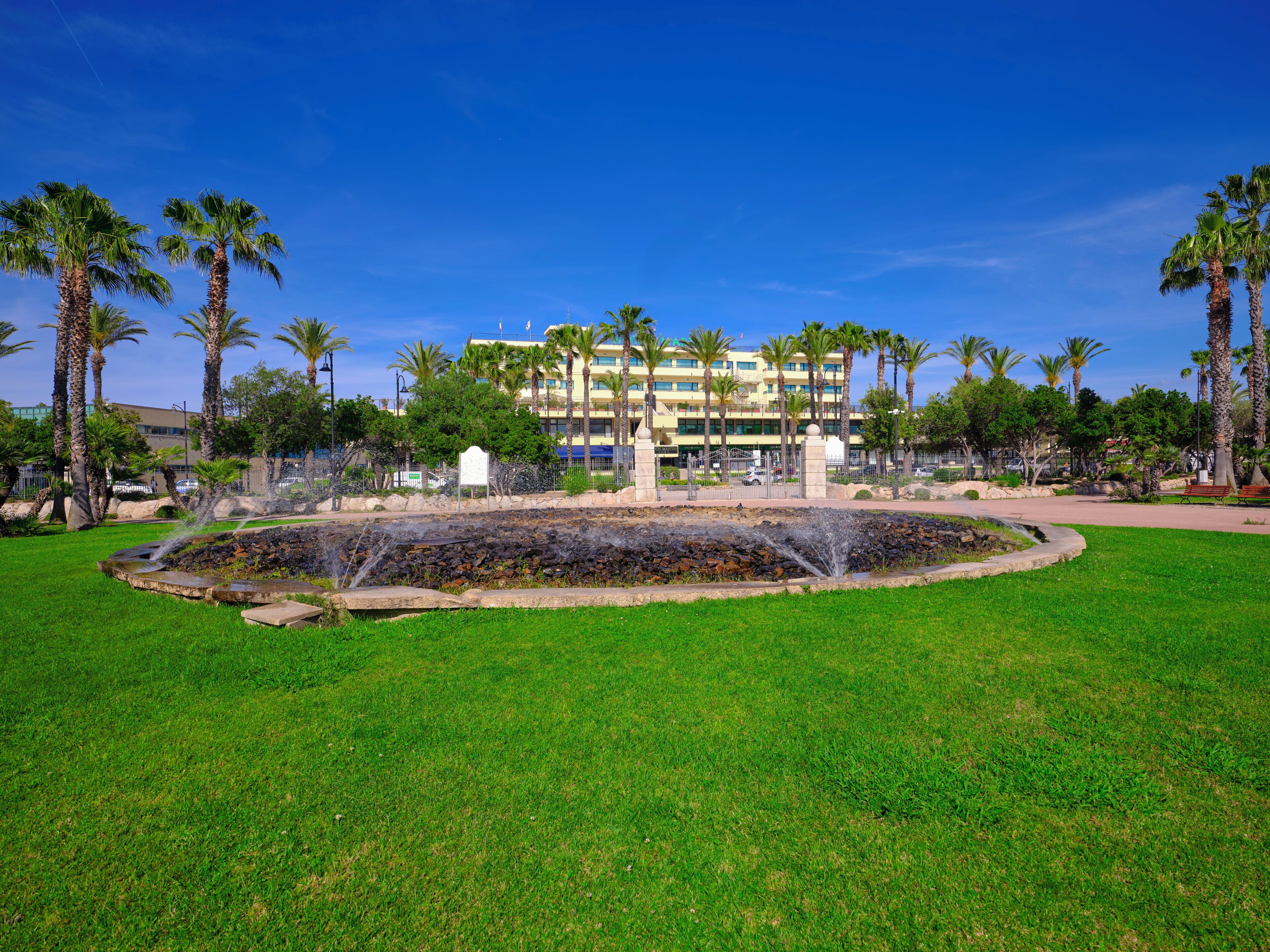 A landscaped area with a fountain, palm trees, and a building in the background.