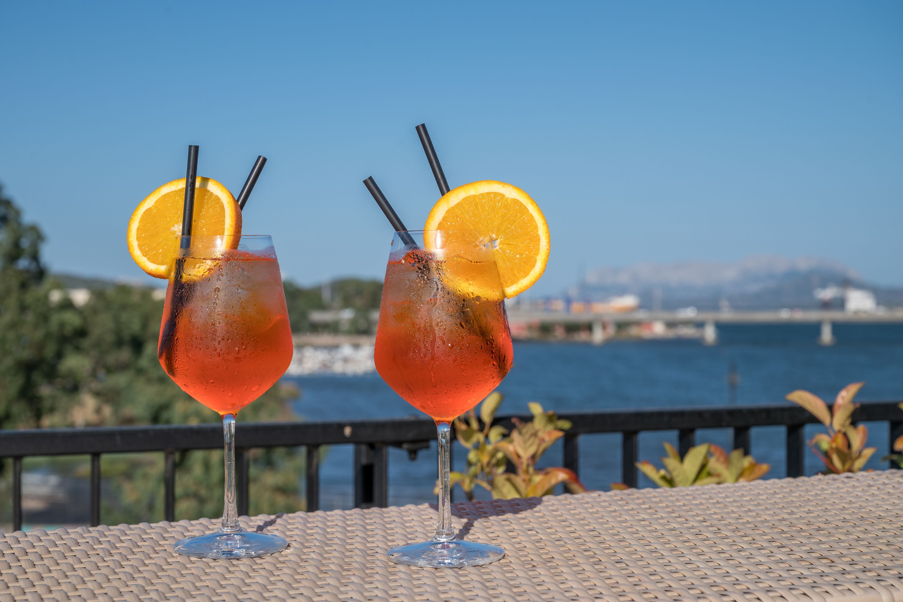 Dos cócteles con rodajas de naranja y pajitas sobre una mesa, con vista al agua y un puente al fondo.