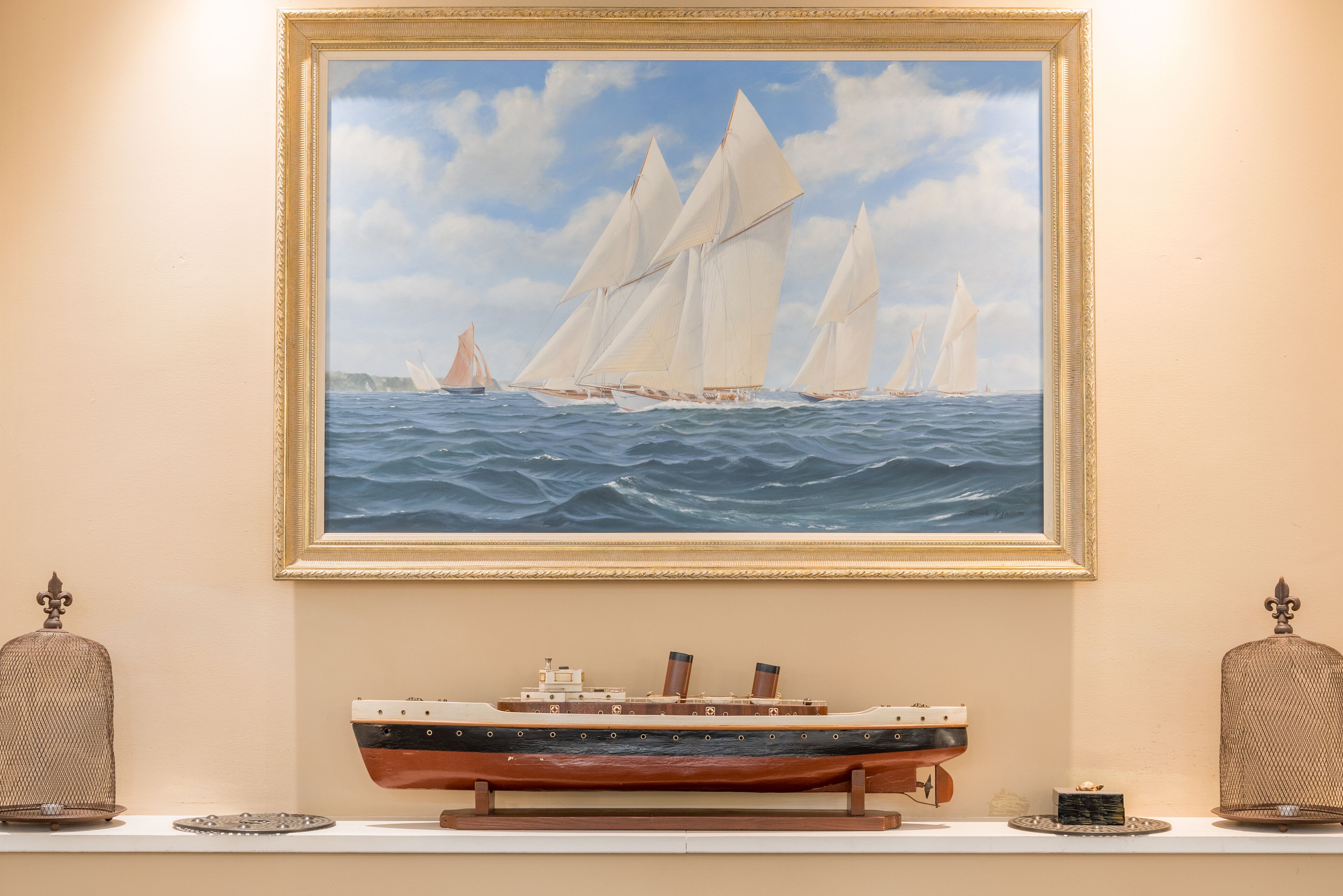 A framed painting of sailboats on the sea, a model ship, and two decorative birdcages on a shelf.