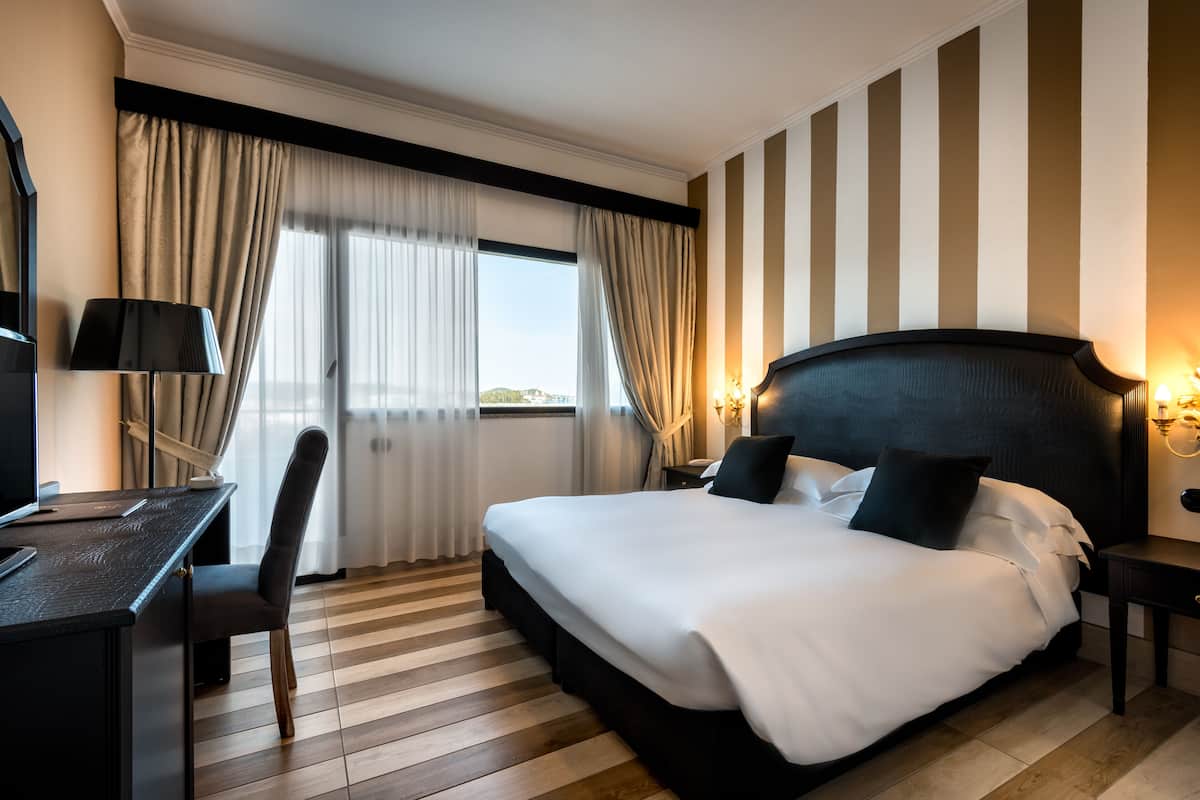 A hotel room with a large bed, a desk with a chair, a computer, and a view of the outdoors through the window.