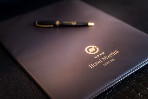 Una libreta de cuero de color oscuro con una pluma encima, que presenta el texto “Hotel Martini” y “OLBA (SS)” grabado en la cubierta.