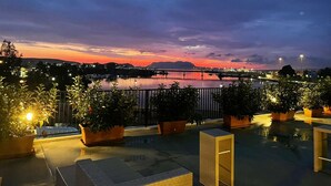 A rooftop terrace with potted plants, outdoor seating, and a view of a river at sunset.