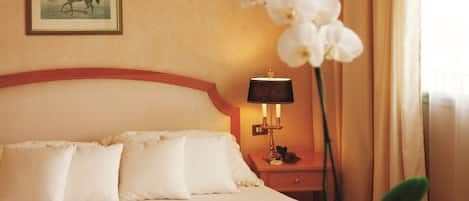 A hotel room with a bed, pillows, a nightstand with a lamp, a framed picture on the wall, and an orchid plant.